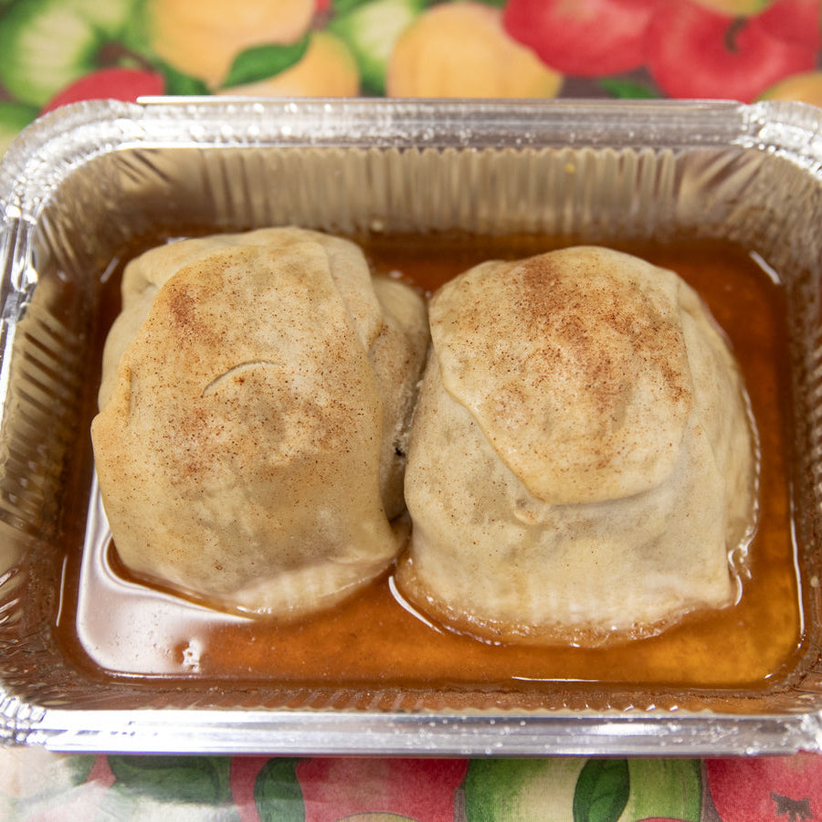 Two apple dumplings in a foil baking pan with a brown sugar glaze on top, on a table with a fruit patterned tablecloth.