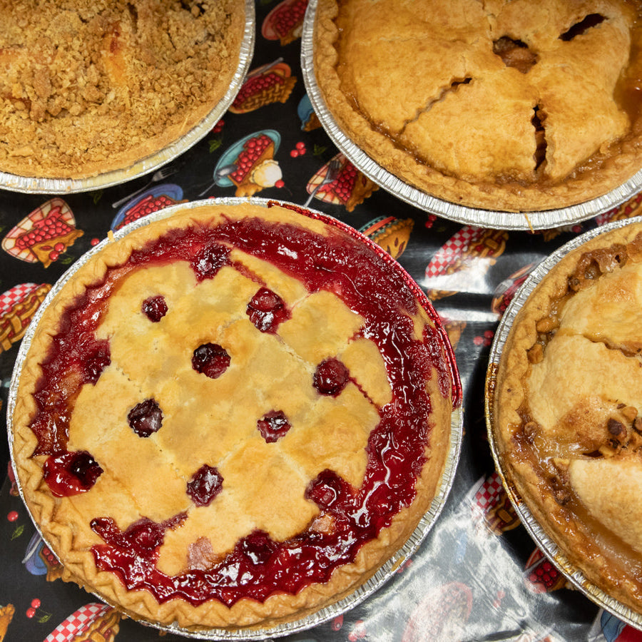 Assortment of freshly baked pies displayed on a table, including a cherry pie with a lattice crust, an apple pie with a solid crust, and others in the background.