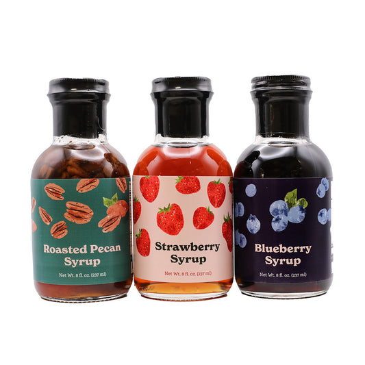 Three bottles of syrup with labels for Roasted Pecan, Strawberry, and Blueberry on a white background.